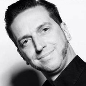 Ein Mann mit kurzen Haaren und Koteletten lächelt leicht in einem Schwarz-Weiß-Porträt und trägt eine dunkle Jacke.