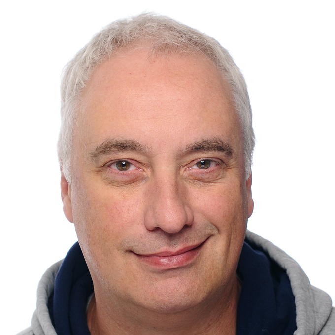 Ein lächelnder Mann mit grauen Haaren vor weißem Hintergrund, trägt eine graue Kapuzenjacke.