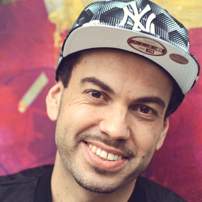 Ein Mann mit Kappe lächelt vor einer bunten, abstrakten Wand.