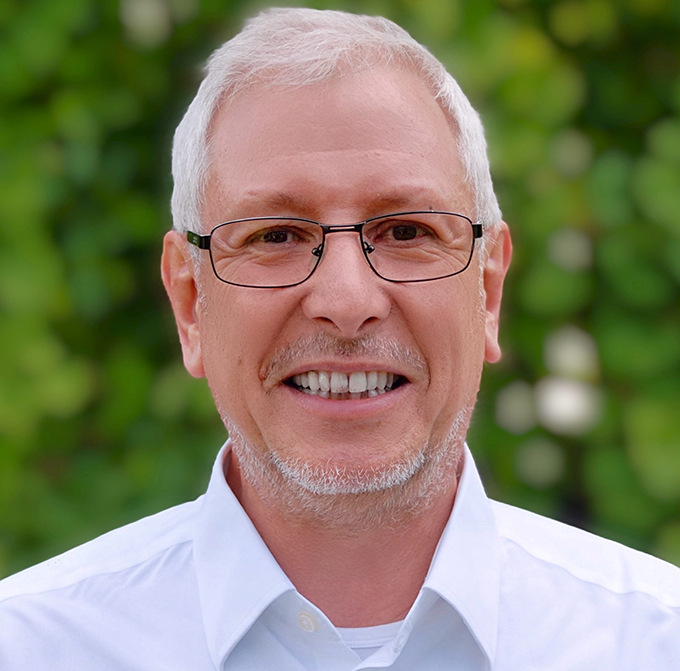 Ein lächelnder Mann mit Brille und weißem Haar vor unscharfem, grünem Hintergrund.