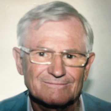 Ein älterer Mann mit Brille lächelt leicht, trägt einen grauen Anzug vor einem neutralen Hintergrund.