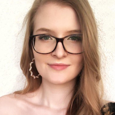 Eine Frau mit Brille und langen Haaren vor hellem Hintergrund, trägt ein offenes Oberteil und große Ohrringe.