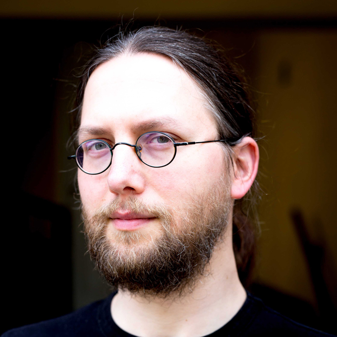 Ein Mann mit Brille und Bart blickt ernst in die Kamera, Hintergrund unscharf.