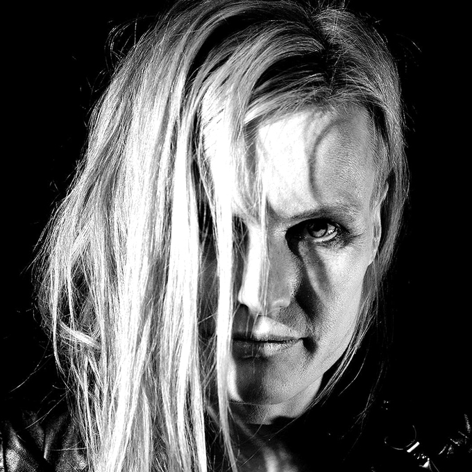 Schwarz-weiß Porträt einer Person mit hellem, langem Haar, das halb das Gesicht bedeckt, vor dunklem Hintergrund.