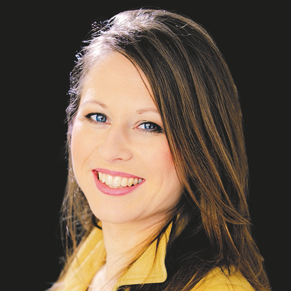 Eine lächelnde Frau mit langen Haaren und gelber Jacke vor schwarzem Hintergrund.