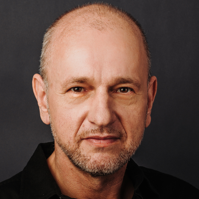 Ein Mann mit Glatze und Bart blickt ernst in die Kamera vor einem dunklen Hintergrund.