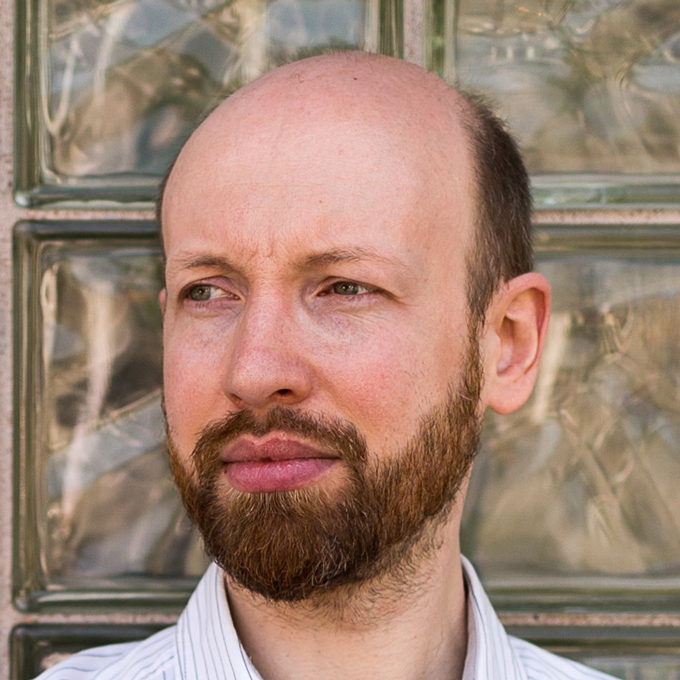 Ein Mann mit Bart und Glatze blickt nach links vor einer Wand aus Glasbausteinen.