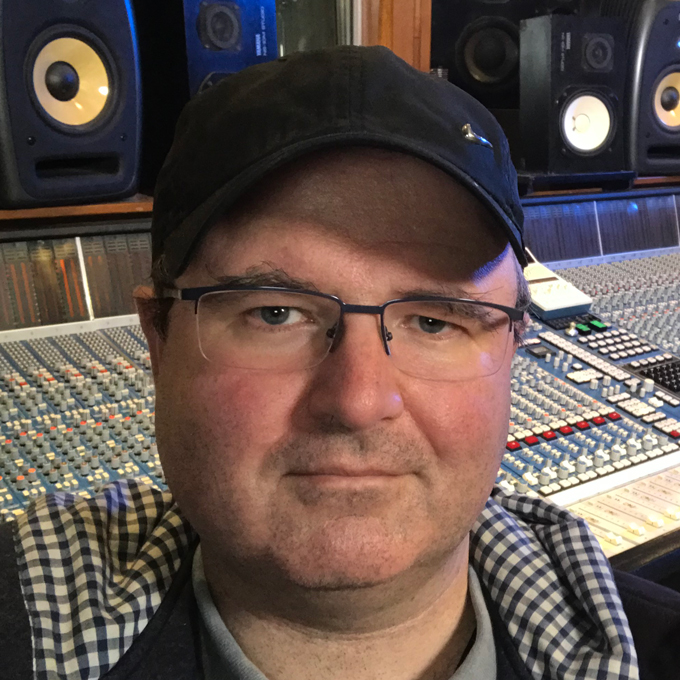 Eine Person mit Brille und Mütze vor einem großen Mischpult im Tonstudio.