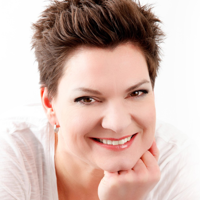 Eine lächelnde Frau mit kurzem braunem Haar stützt ihr Kinn auf die Hand, vor weißem Hintergrund.