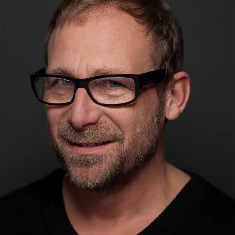 Ein Mann mit Brille lächelt vor einem dunklen Hintergrund.