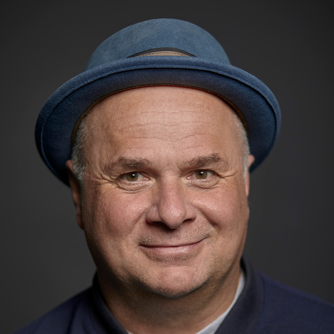 Ein lächelnder Mann mit blauem Hut vor grauem Hintergrund.