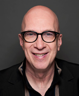 Eine Person mit Brille lächelt vor einem grauen Hintergrund.