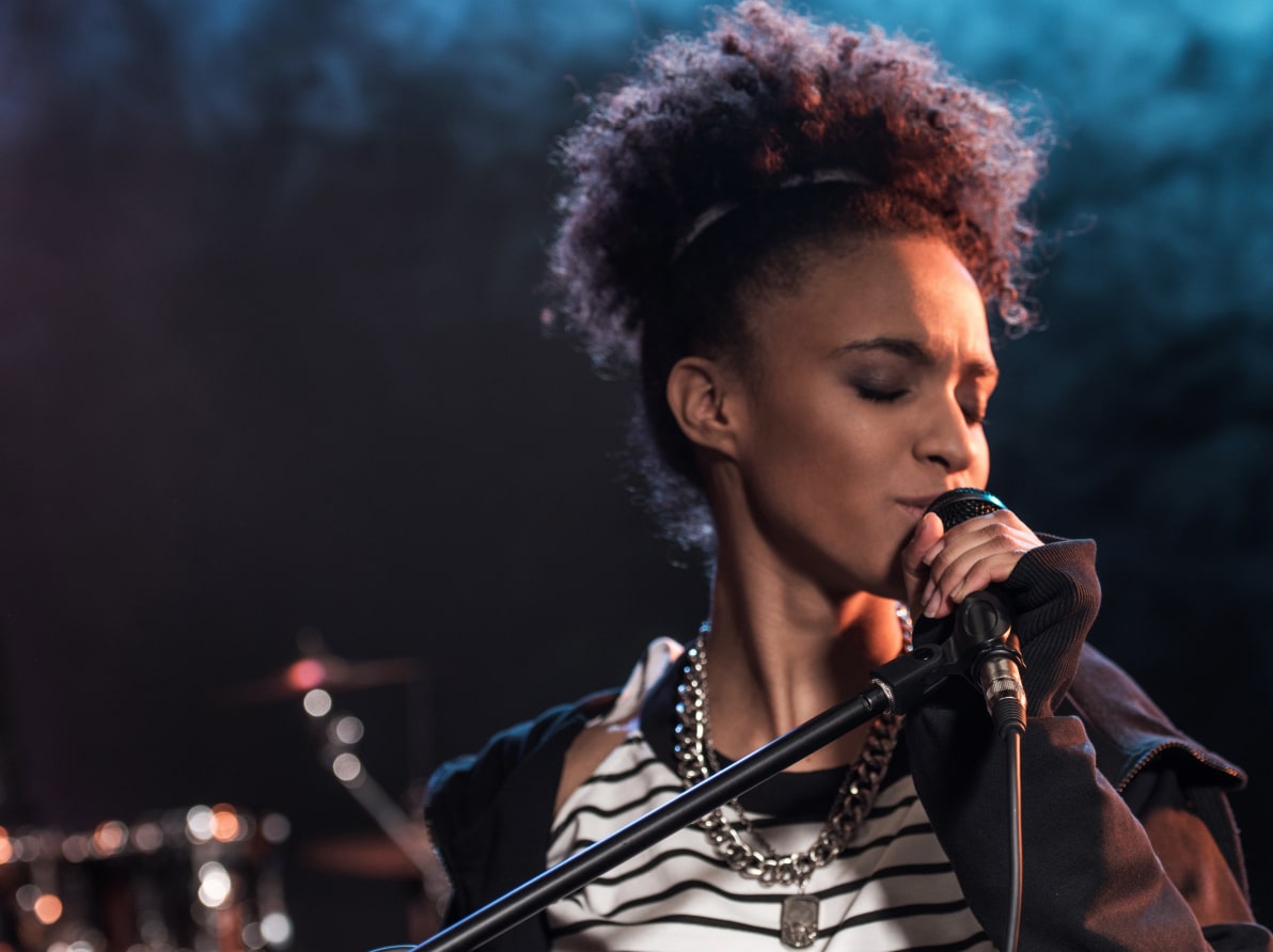 Eine Frau singt leidenschaftlich in ein Mikrofon auf einer Bühne, im Hintergrund verschwommene Schlagzeug-Elemente.