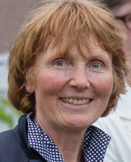 Eine Person mit kurzen, hellbraunen Haaren lächelt im Freien, sie trägt eine dunkle Jacke und ein gepunktetes Hemd.