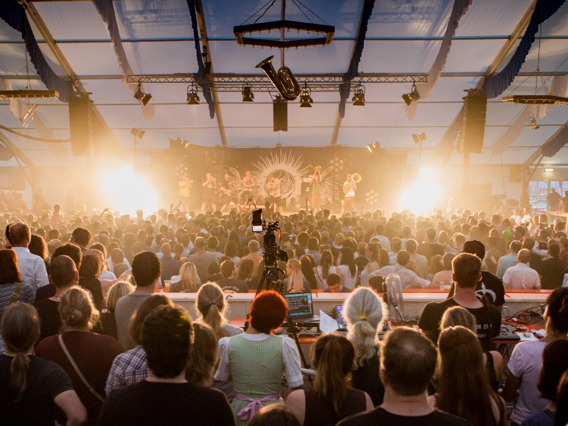 Eine große Menschenmenge in einem Festzelt schaut auf eine beleuchtete Bühne mit Musikern.