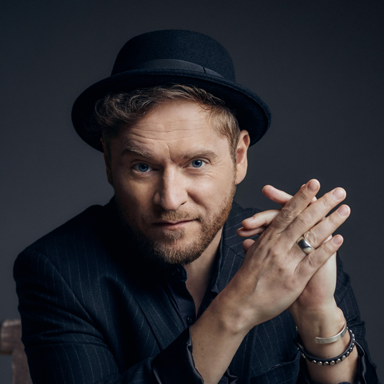 Mann mit Bart und Hut in dunklem Anzug, Hände gefaltet, blickt direkt in die Kamera.