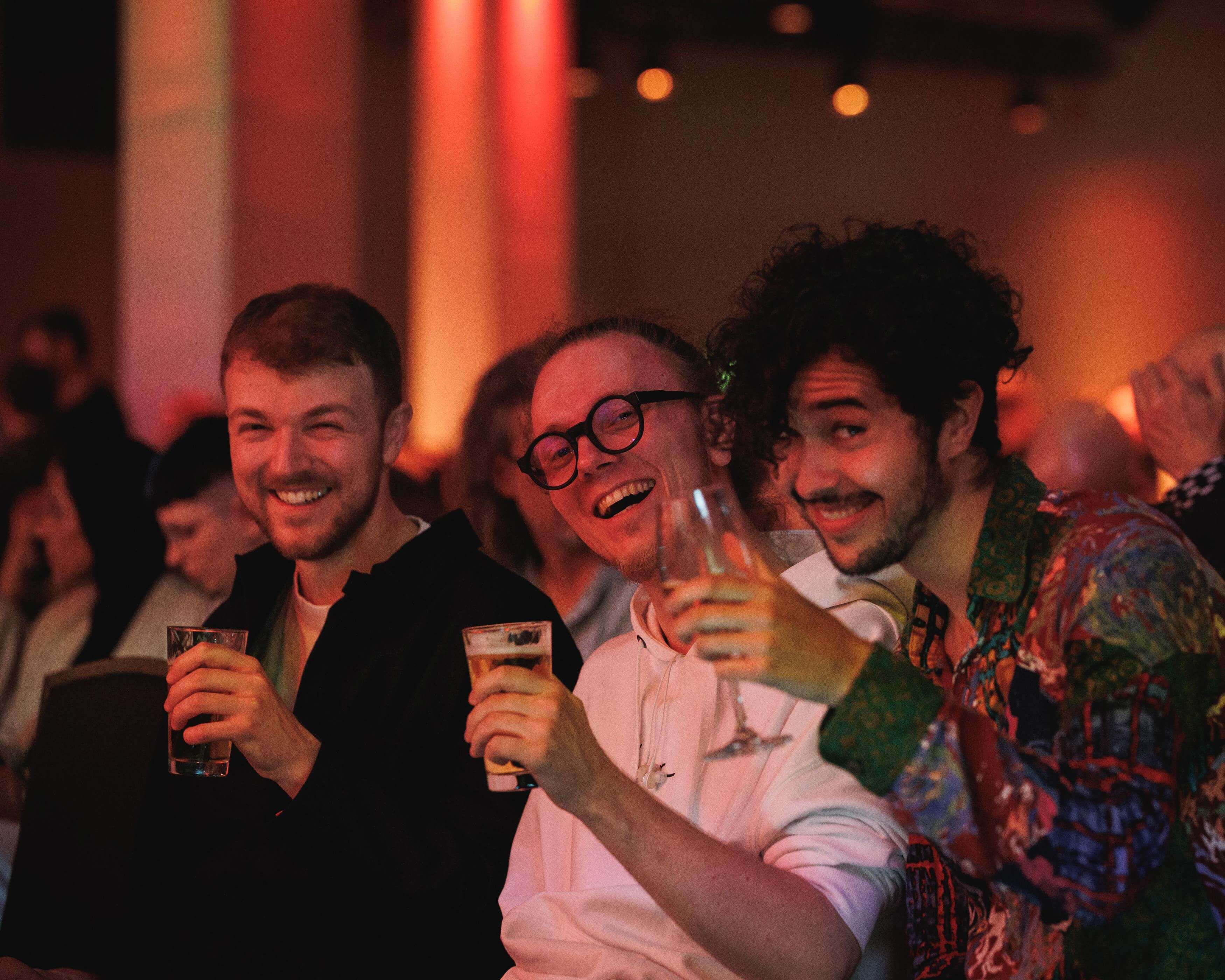 Drei lachende Männer halten Gläser in einer bunt beleuchteten Menschenmenge.