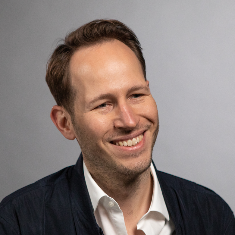 Ein lächelnder Mann in einem weißen Hemd und einer dunklen Jacke vor einem schlichten grauen Hintergrund.