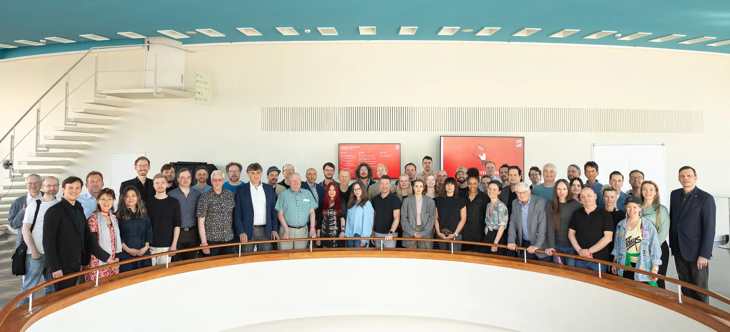 Eine Gruppe von Menschen steht in einem modernen Raum vor einer weißen Wand mit zwei roten Tafeln.