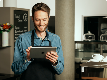 Ein Mann steht mit einer schwarzen Schürze in einem Café und schaut dabei auf ein iPad.