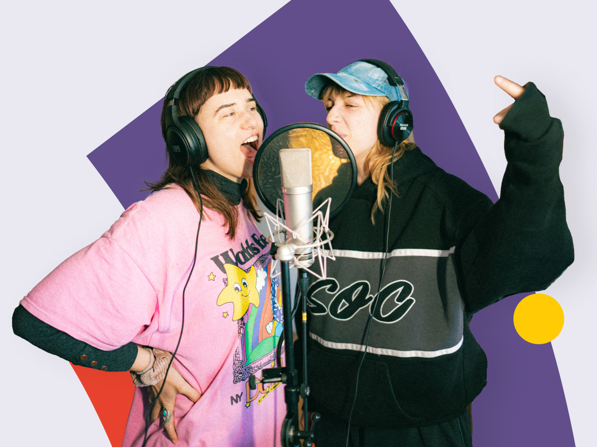 Zwei Personen singen in ein Studiomikrofon, tragen Kopfhörer, vor einem bunten Hintergrund.