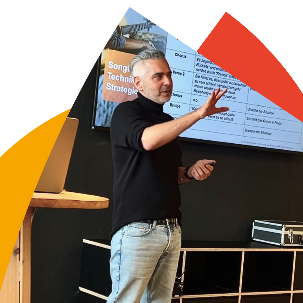 Ein Mann in schwarzem Pullover präsentiert vor einem Bildschirm mit Text und Grafiken in einem Raum.