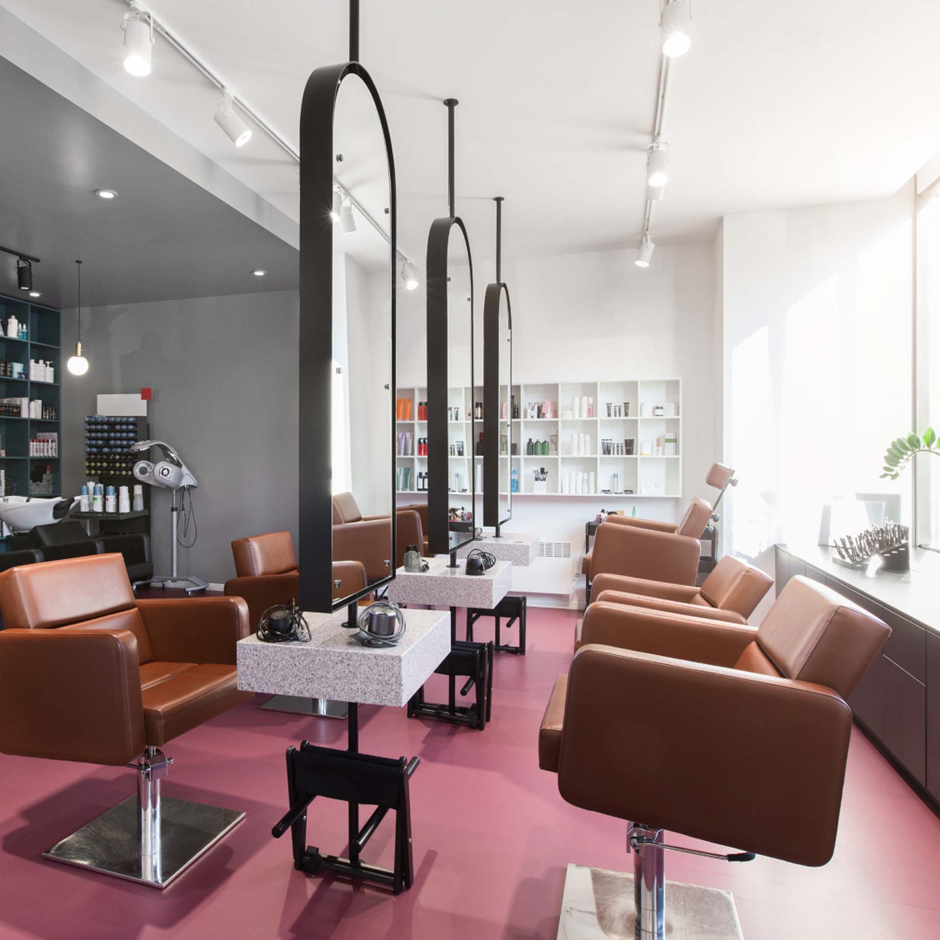 Moderner Friseursalon mit braunen Stühlen, großen Spiegeln und Regalen voller Haarprodukte auf rosa Boden.