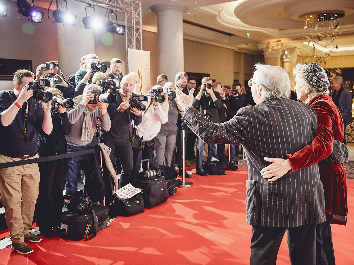 Ein älteres Paar posiert auf rotem Teppich vor Fotografen mit Kameras.