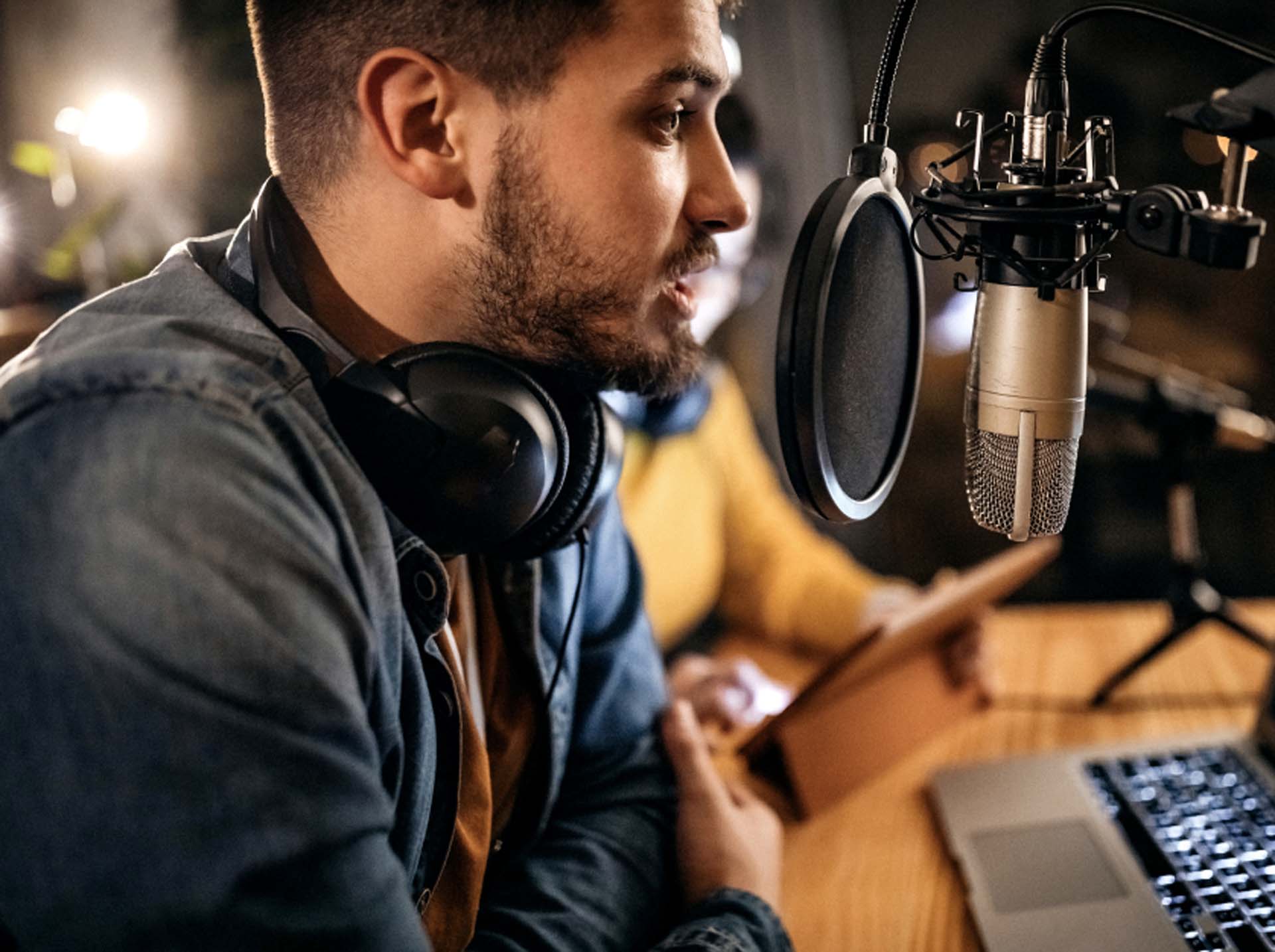 Mann mit Kopfhörern spricht in ein Mikrofon vor einem Laptop.