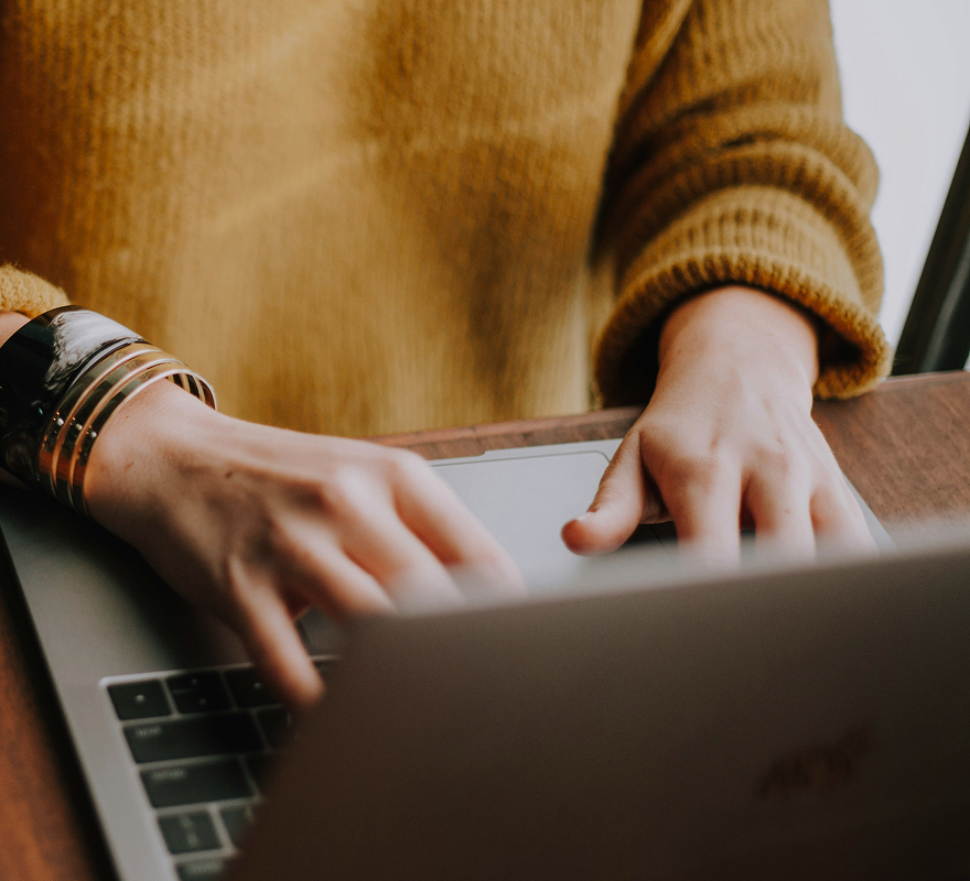 Hände tippen auf einem Laptop, Person trägt gelben Pullover und Armreif.
