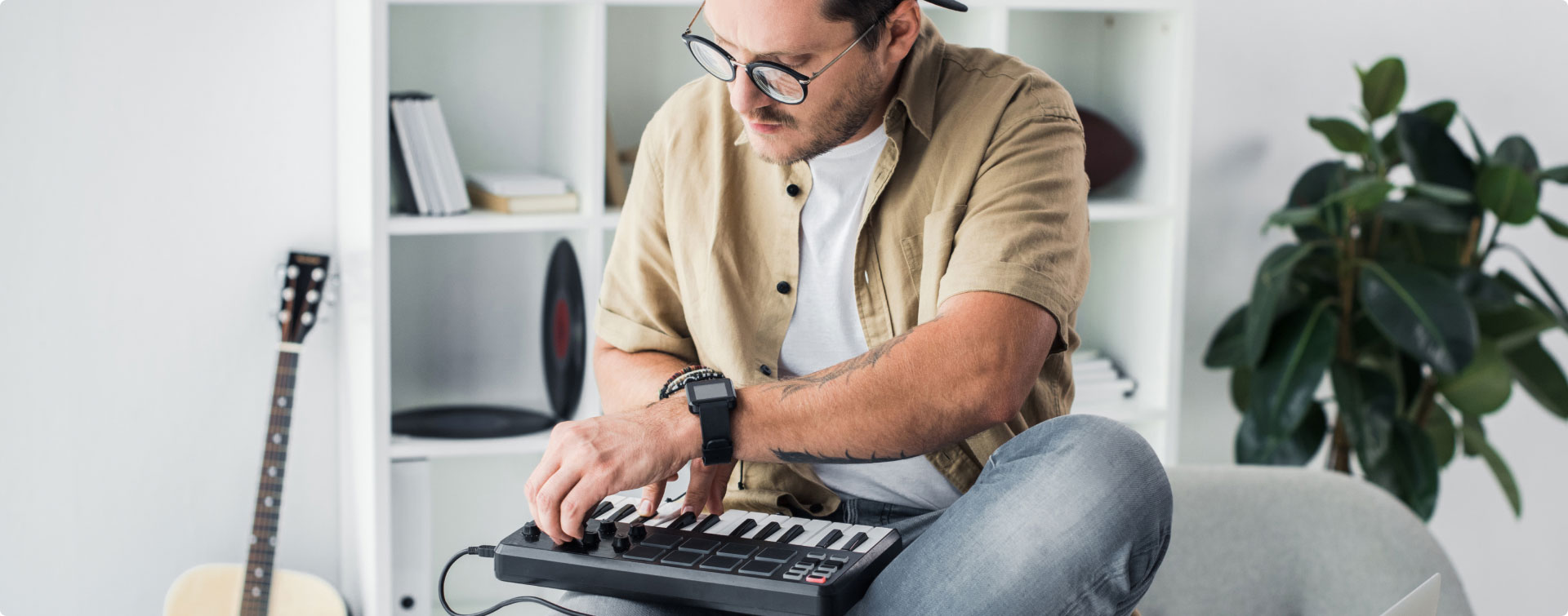 Ein Mann mit Brille spielt ein MIDI-Keyboard, im Hintergrund eine Gitarre und ein Regal.