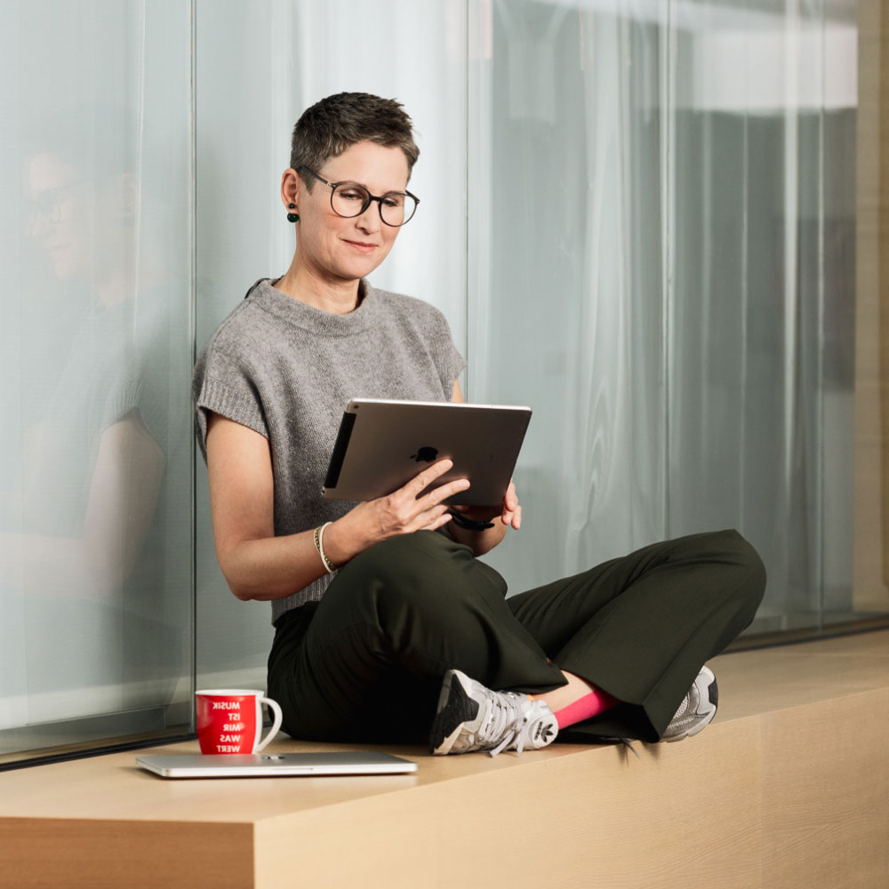 Person mit Brille sitzt auf Fensterbank, hält Tablet, neben roter Tasse und Laptop.