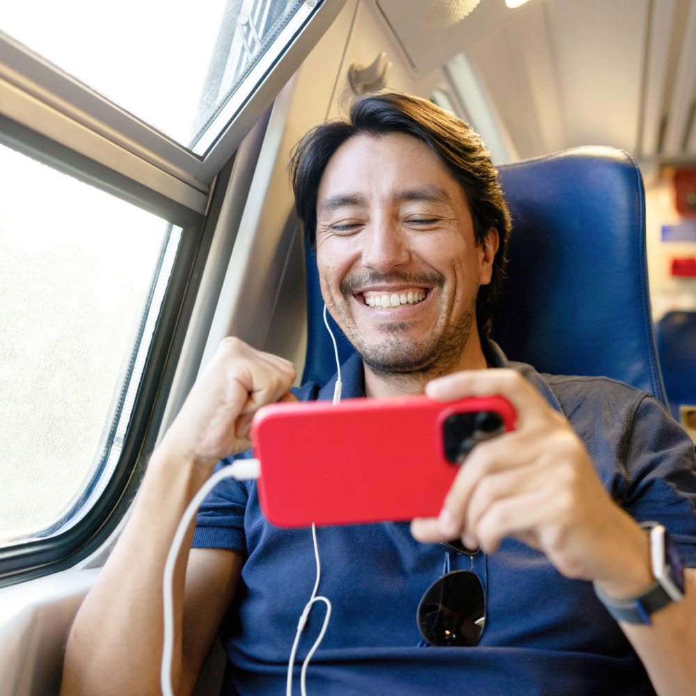 Ein Mann im Zug lächelt, während er auf ein rotes Smartphone schaut und Kopfhörer trägt.