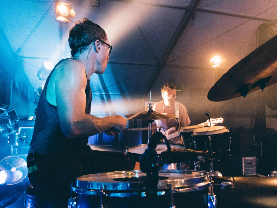 Zwei Schlagzeuger spielen auf einer Bühne, umgeben von buntem Scheinwerferlicht.