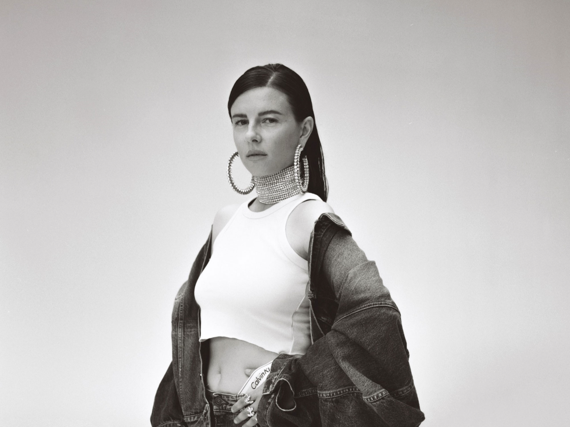 Frau in Jeansjacke und weißem Top, große Ohrringe, schaut ernst in die Kamera vor hellem Hintergrund.