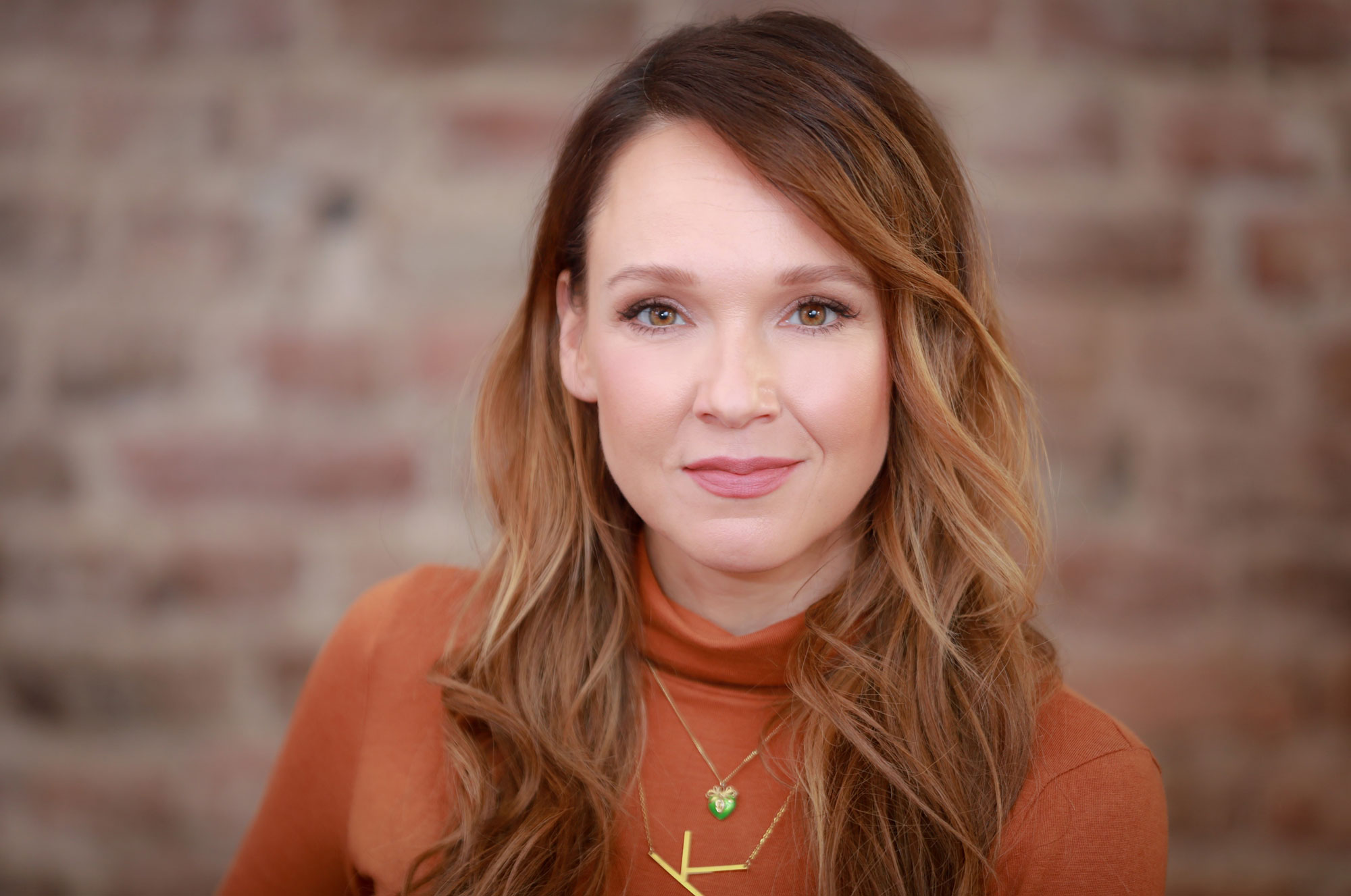 Eine Frau mit langen, braunen Haaren vor einer Backsteinwand, trägt ein braunes Oberteil und zwei Halsketten.