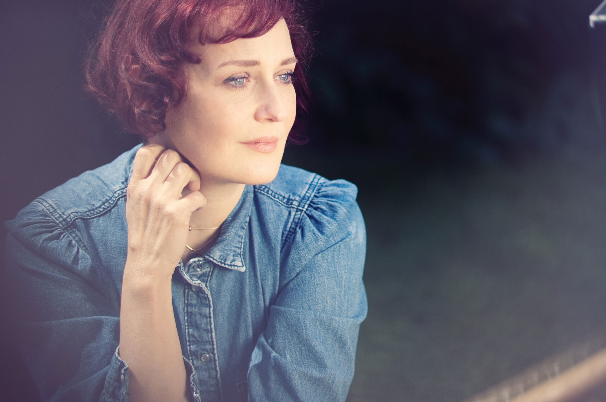 Eine Frau mit roten Haaren und Jeanshemd schaut nachdenklich aus einem Fenster.