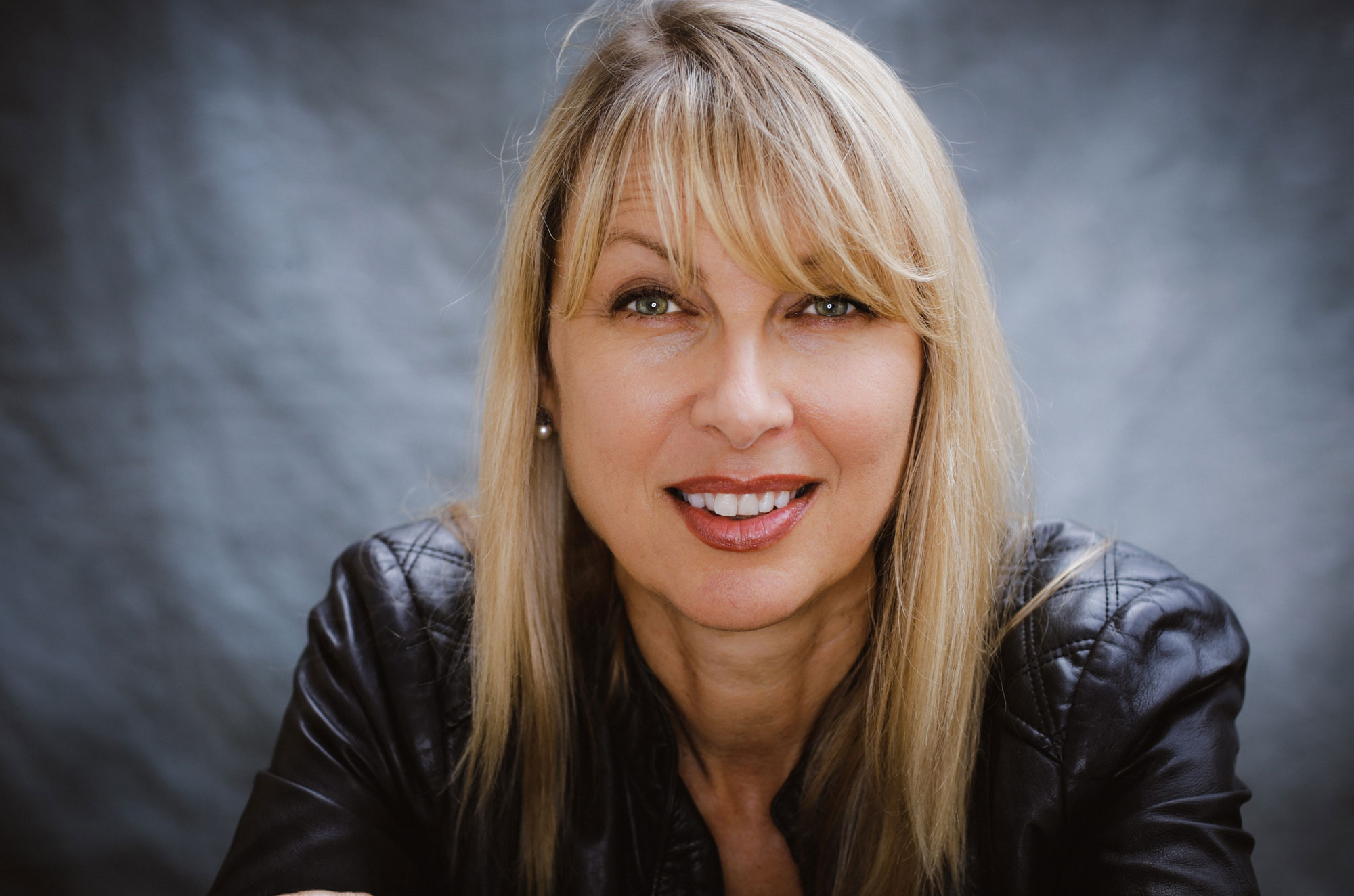 Eine Frau mit blonden Haaren lächelt in einer schwarzen Lederjacke vor grauem Hintergrund.