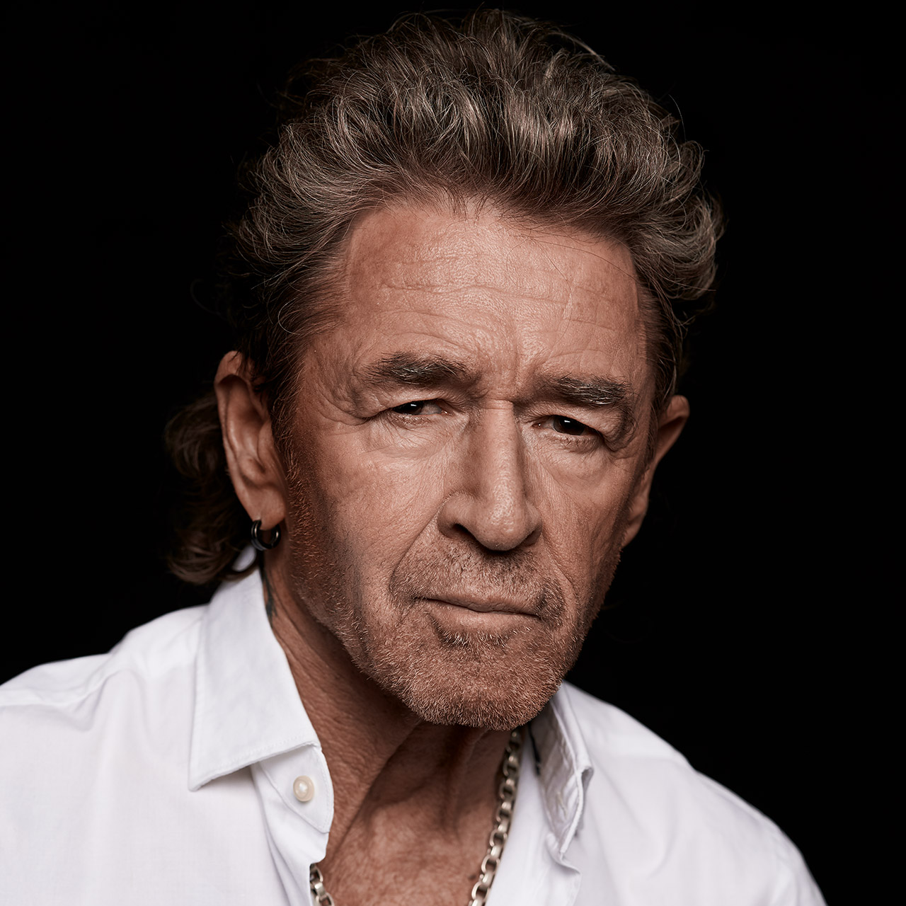 Ein älterer Mann mit grauem Haar und Bart trägt ein weißes Hemd vor schwarzem Hintergrund.