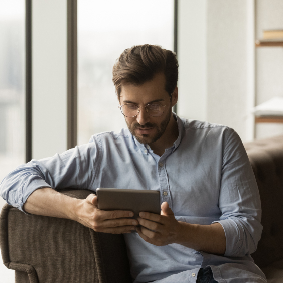 Ein Mann mit Brille liest auf einem Tablet, sitzt auf einem Sofa vor einem großen Fenster.