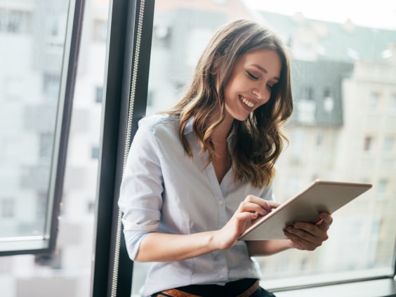 Eine lächelnde Frau sitzt am Fenster und benutzt ein Tablet.