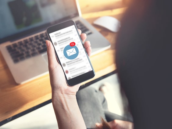 Eine Hand hält ein Smartphone mit 12 neuen E-Mails vor einem Laptop.