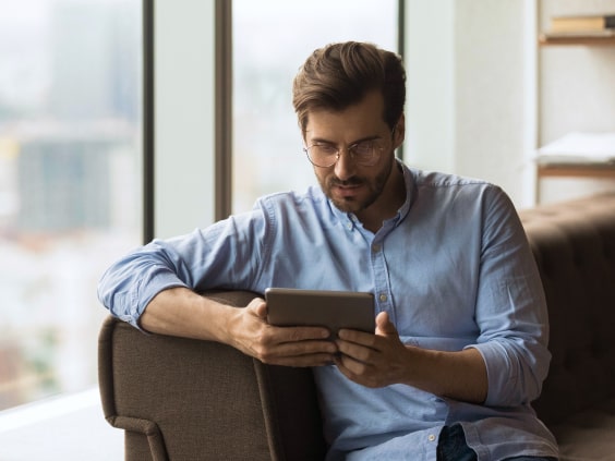 Ein Mann mit Brille liest ein Tablet auf einem Sofa vor einem großen Fenster.