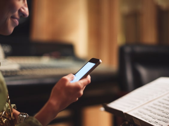 Person hält Smartphone in der Hand, daneben ein Notenständer mit Notenblättern, im Hintergrund ein unscharfes Mischpult.