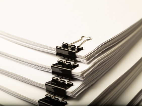 Ein Stapel weißer Papiere, zusammengehalten von schwarzen Büroklammern.