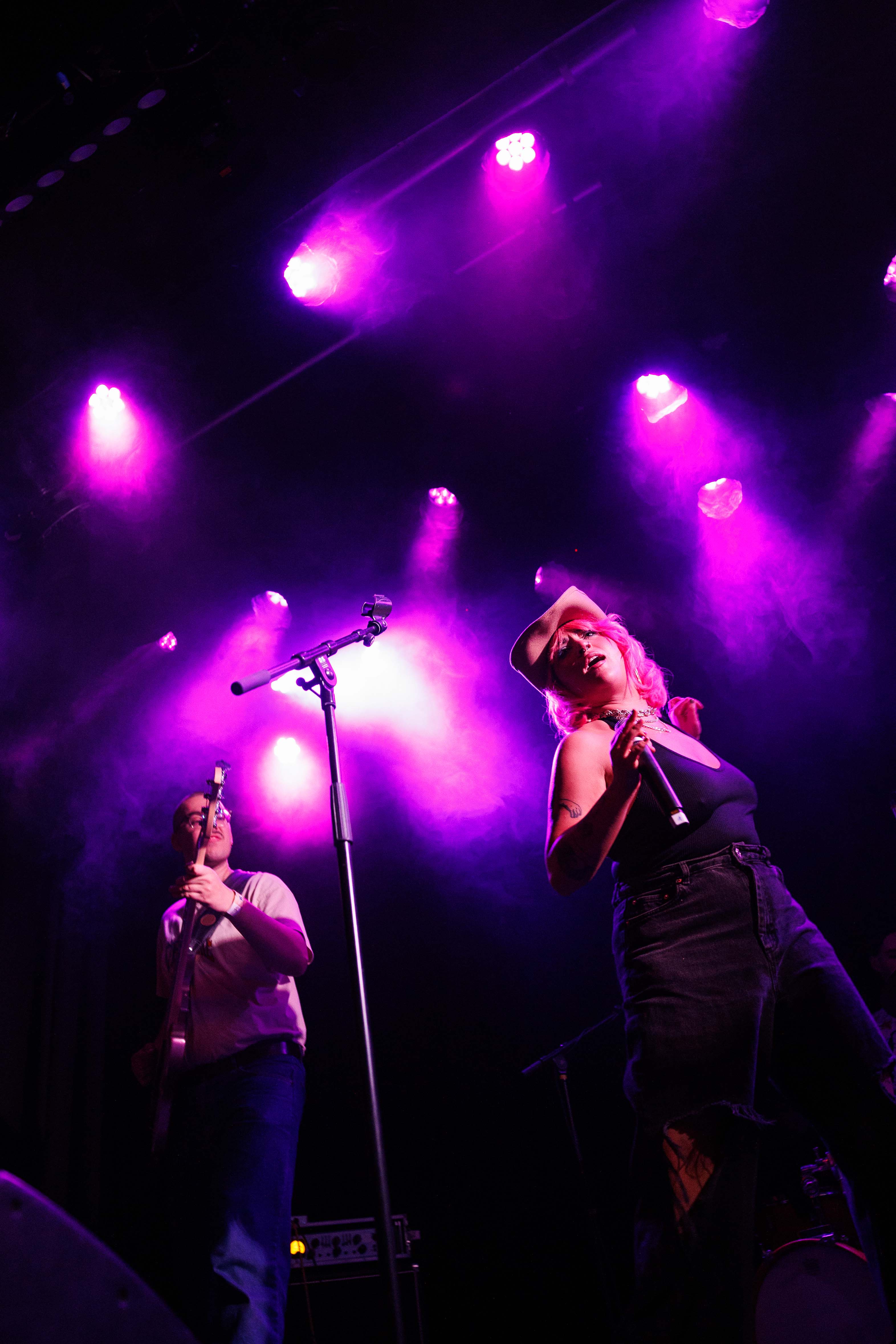 Zwei Musiker auf der Bühne unter lila Scheinwerfern, einer mit Gitarre, eine singend mit Hut.