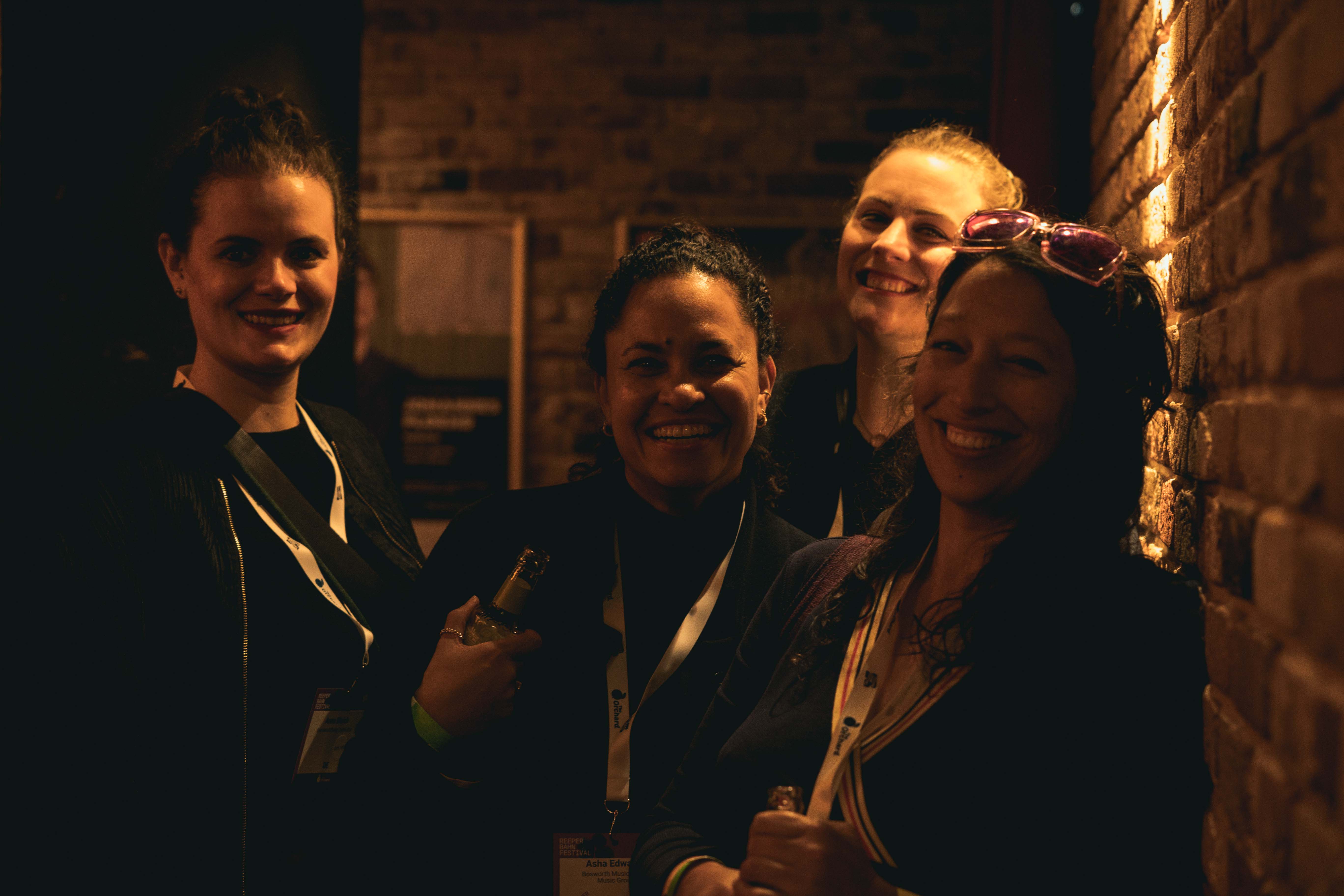 Vier lächelnde Personen stehen in einem schummrigen Raum vor einer Backsteinwand.