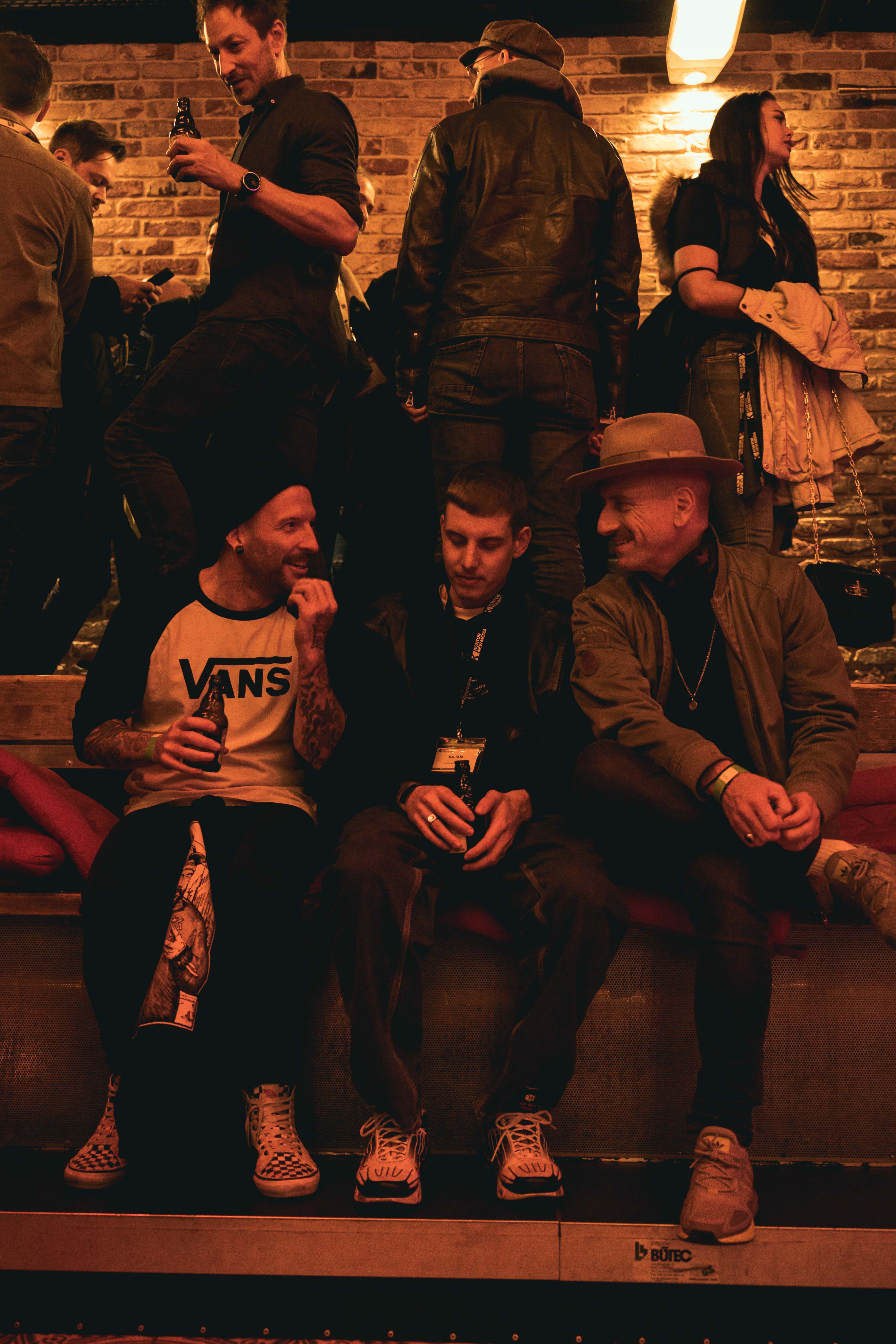 Drei Männer sitzen auf einer Bank in einer Bar, umgeben von stehenden Personen, mit Getränken in der Hand.