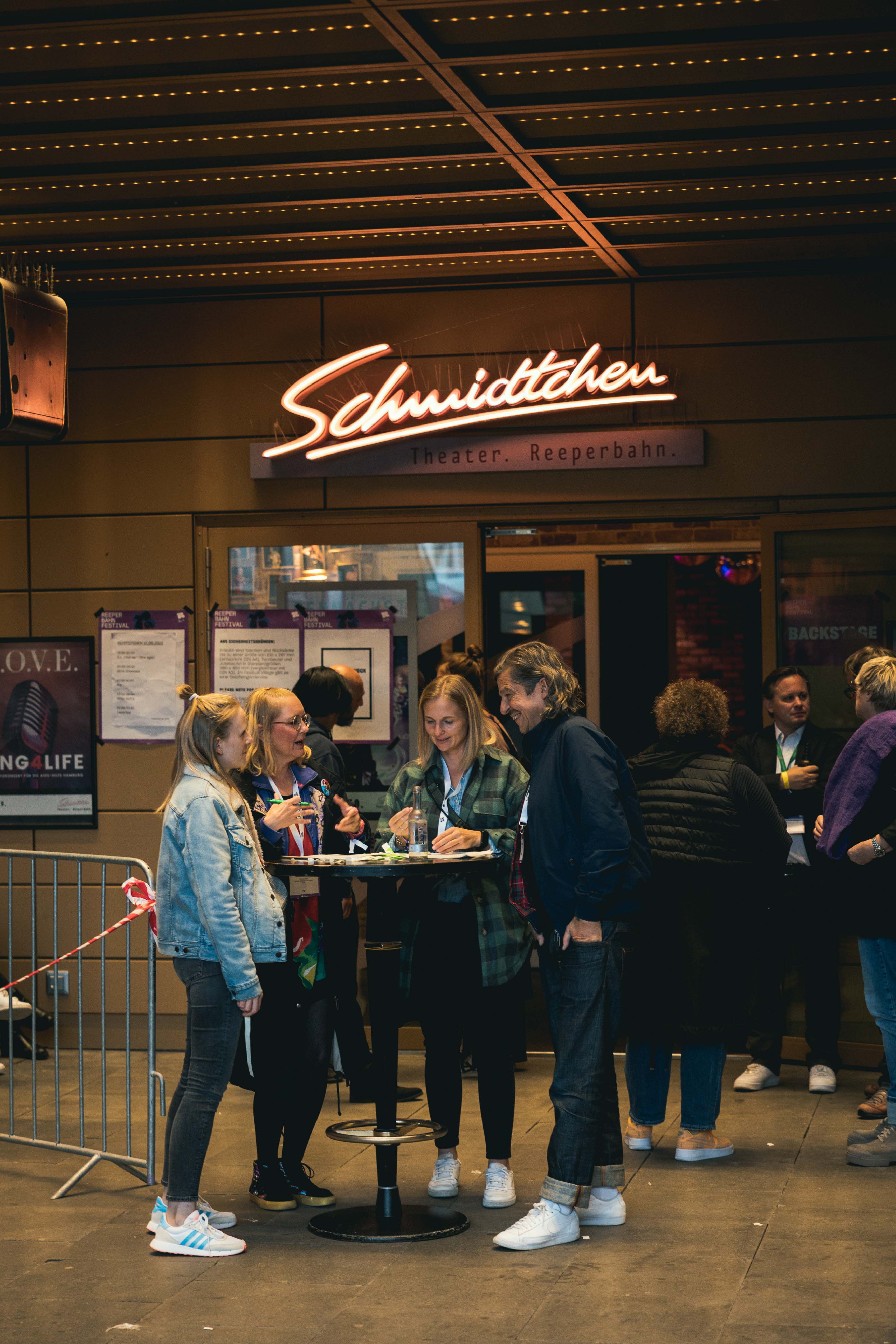 Personen stehen vor dem Schmidtchen Theater unter einem beleuchteten Schild und unterhalten sich an einem Stehtisch.