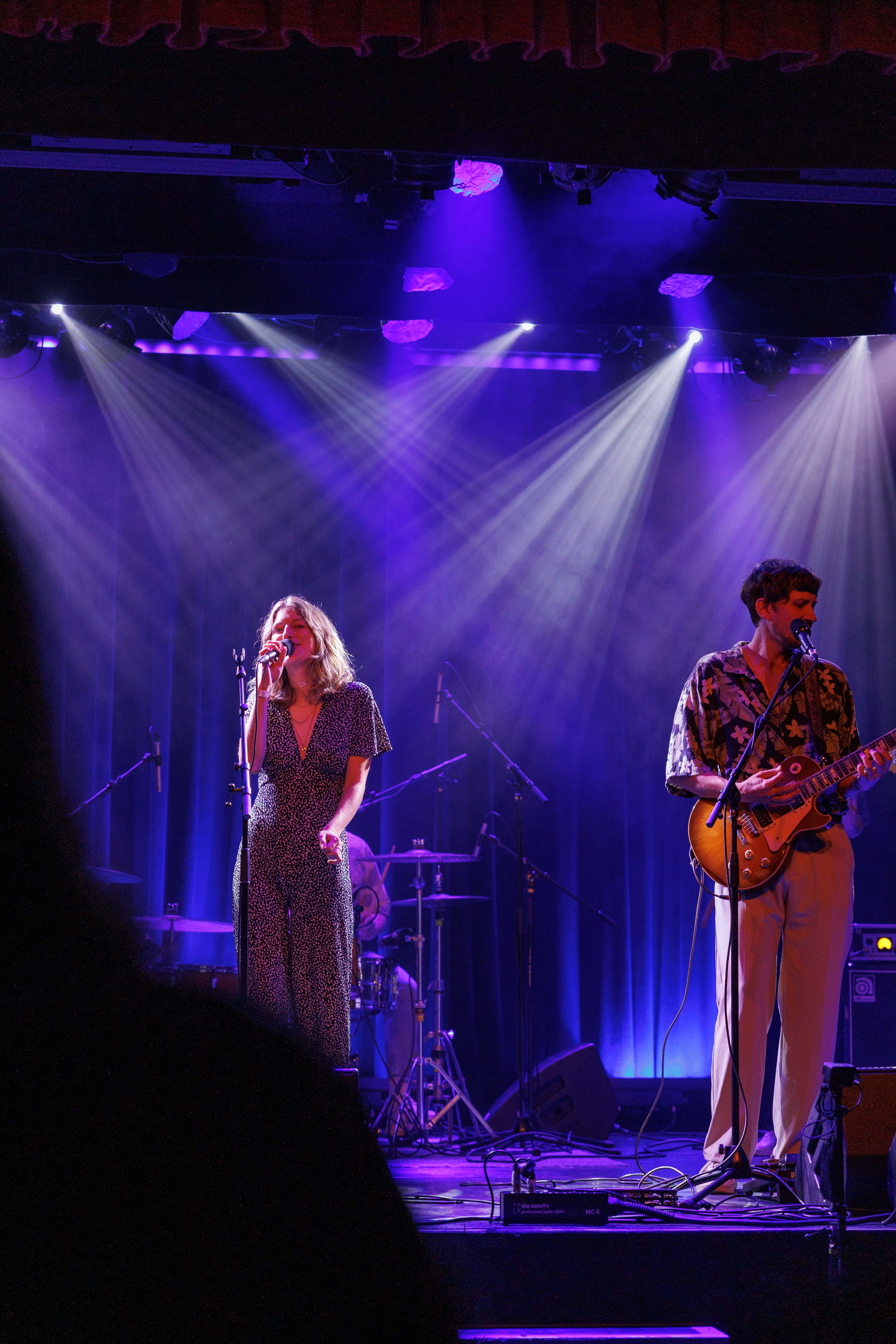 Sängerin und Gitarrist auf einer Bühne in violettem Licht, Mikrofone und Schlagzeug im Hintergrund.